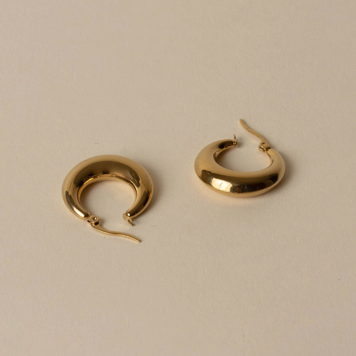 A pair of polished gold crescent-shaped hoop earrings with fishhook backs, displayed on a neutral beige background.
