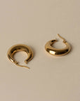 A pair of polished gold crescent-shaped hoop earrings with fishhook backs, displayed on a neutral beige background.