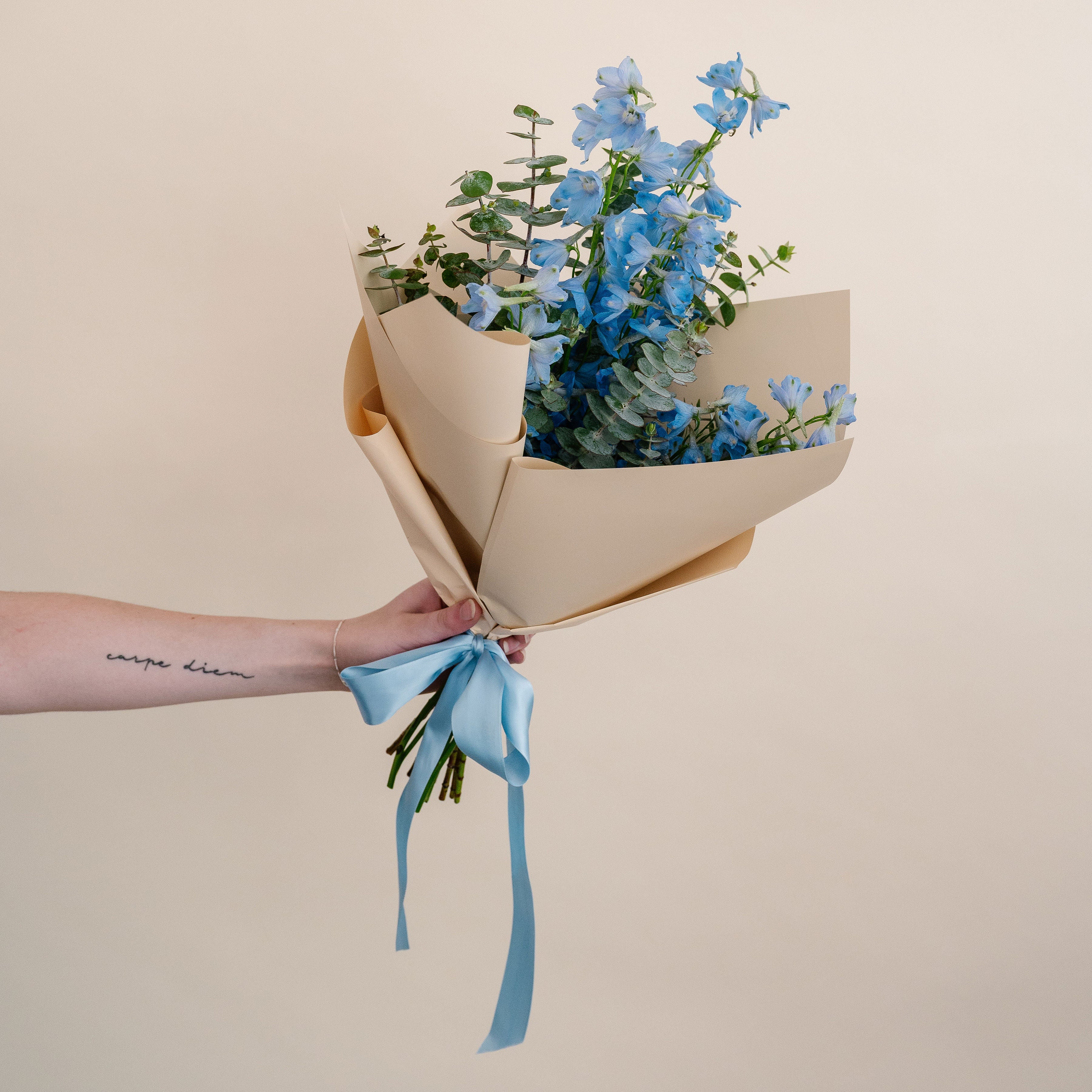 Bouquet of blue flowers wrapped in brown paper with a blue ribbon held by a person against a beige background