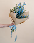 Bouquet of blue flowers wrapped in brown paper with a blue ribbon held by a person against a beige background