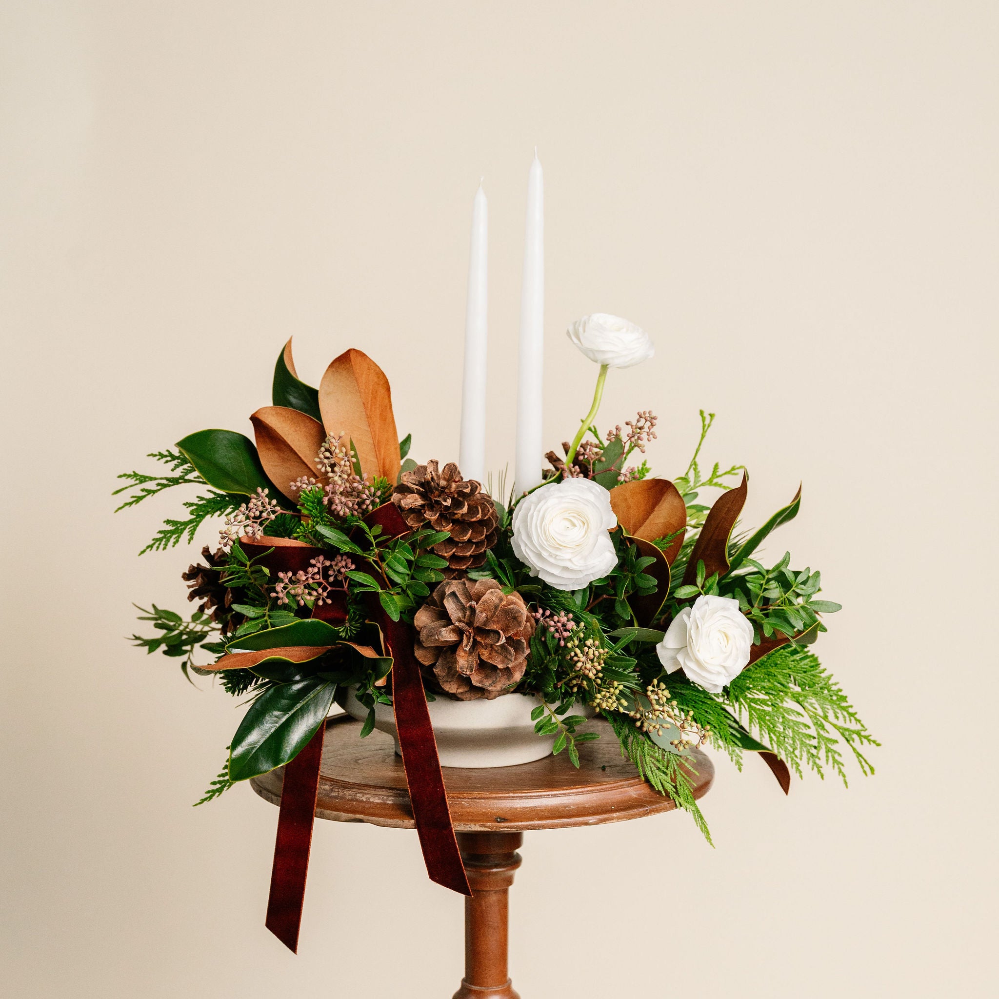 Floral arrangement with pinecones and white candles on a small wooden table against a beige background