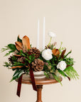 Floral arrangement with pinecones and white candles on a small wooden table against a beige background