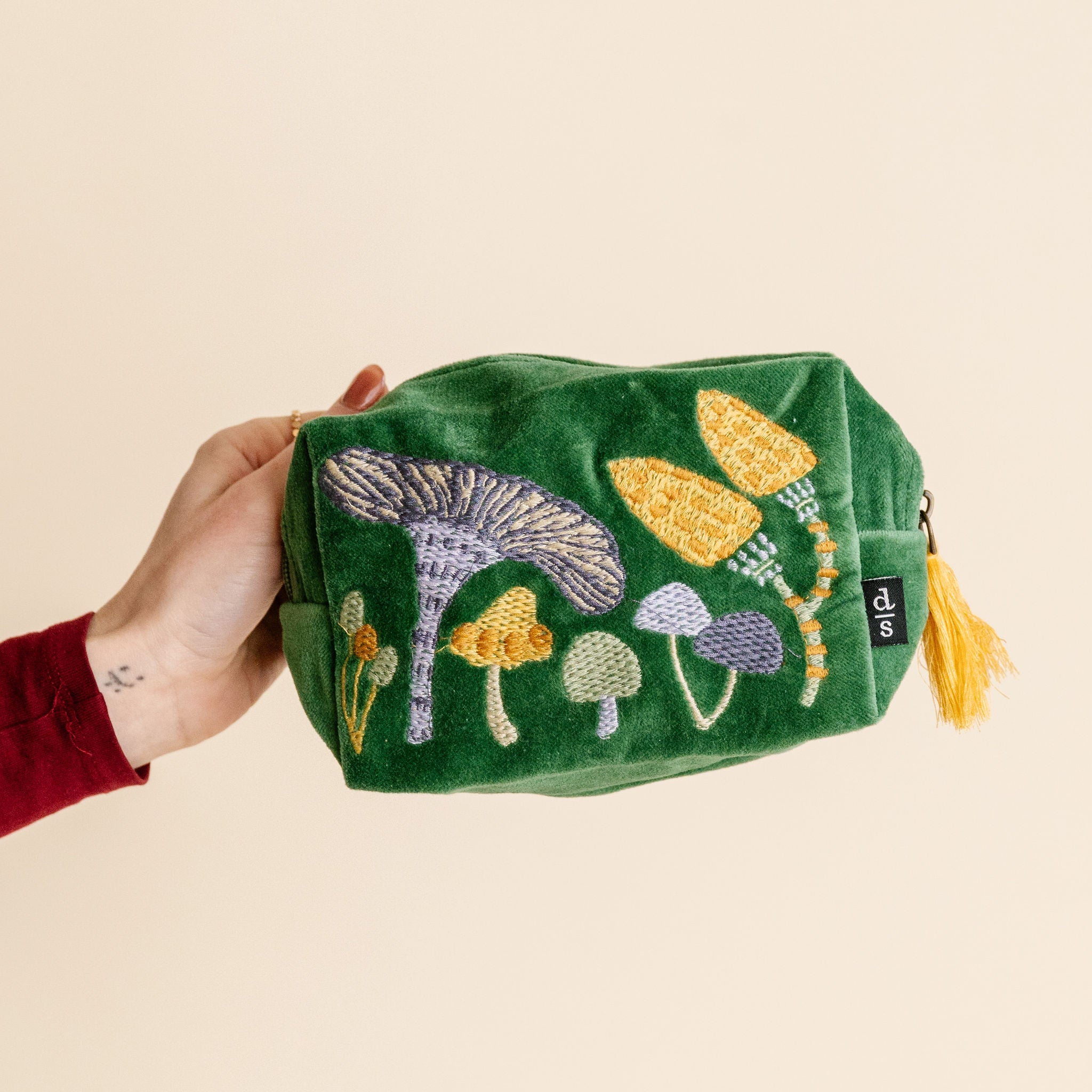 Green pouch with embroidered mushrooms held by a hand on a beige background
