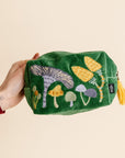 Green pouch with embroidered mushrooms held by a hand on a beige background