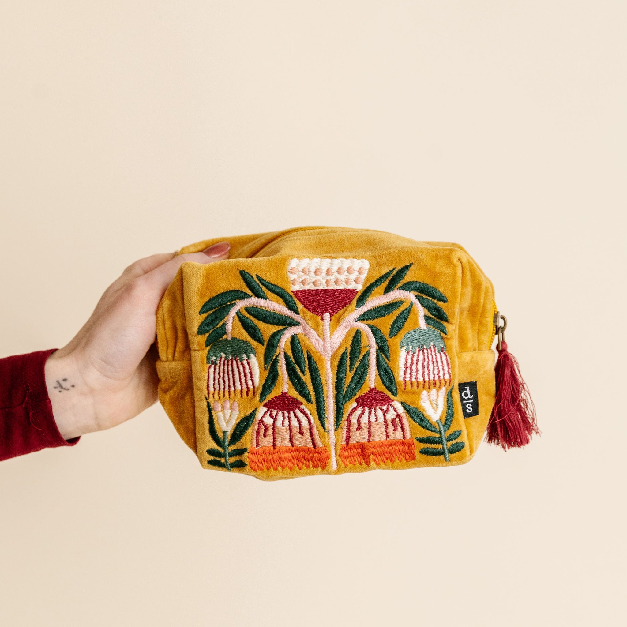 Hand holding a mustard yellow pouch with floral embroidery on a beige background