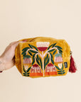 Hand holding a mustard yellow pouch with floral embroidery on a beige background