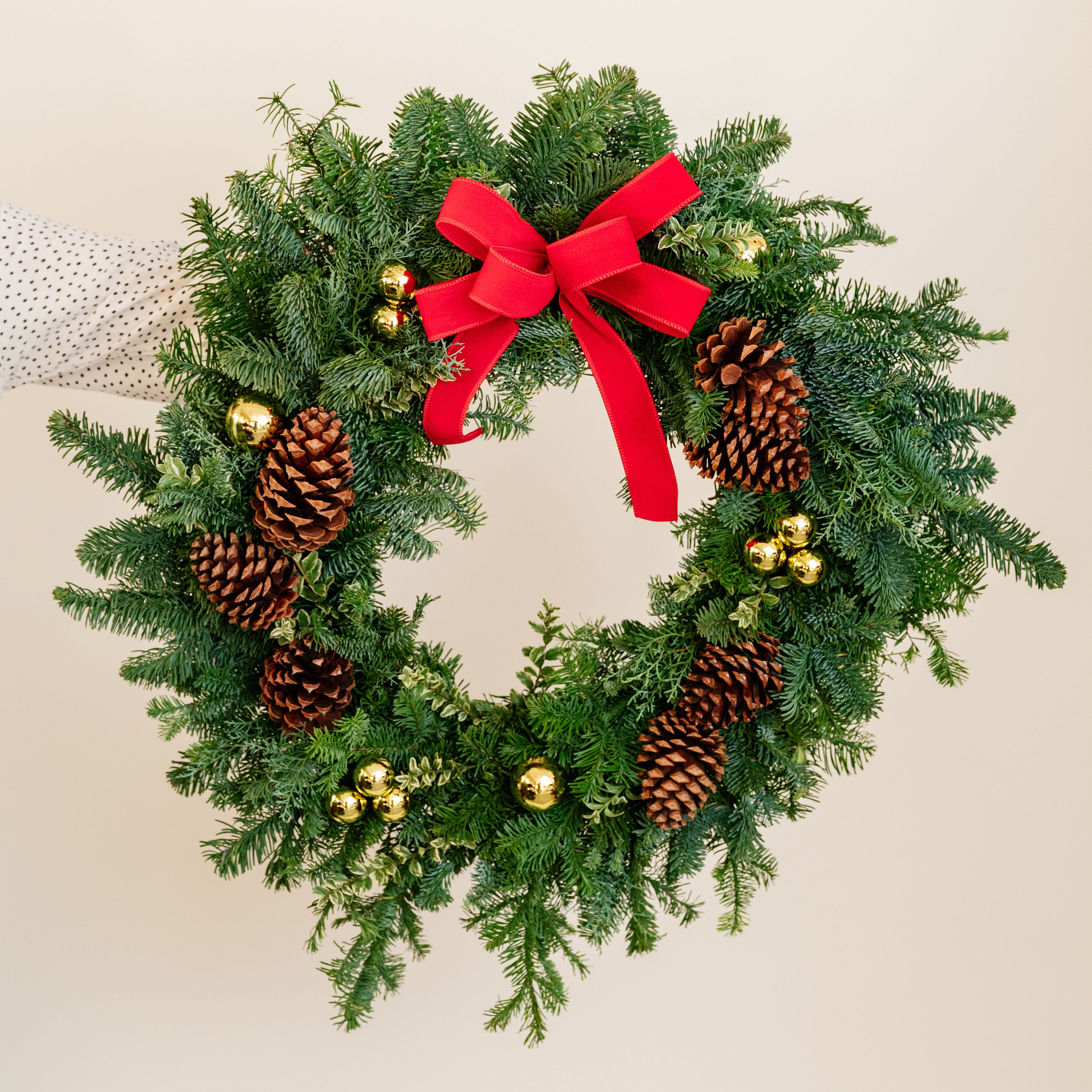 Christmas wreath with greenery, pinecones, gold balls, and a red bow on a neutral background.