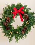 Christmas wreath with greenery, pinecones, gold balls, and a red bow on a neutral background.
