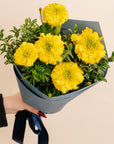Bouquet of yellow flowers wrapped in gray paper held by a person against a beige background
